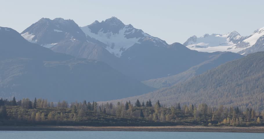 Alaska mountains and glacier patches tower above a forest shoreline as calm water flows across the foreground in a serene wilderness landscape.