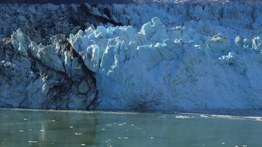 Glacier towers above icy water as the camera tilts upward, revealing blue ice, snow-covered Alaska mountains, and a clear sky from a passing cruise ship.