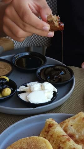 Traditional Moroccan breakfast served with small dishes of amlou, olives, fresh cheese, butter, olive oil, and honey. Close-up of a young man dipping a piece of bread into honey. Authentic Moroccan cuisine, local food culture, and traditional breakfast habits.