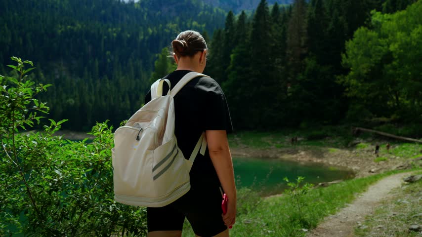 Solo hiker with white backpack walking along tranquil forest lake trail, exploring lush green mountains, peaceful shoreline and calm reflective waters