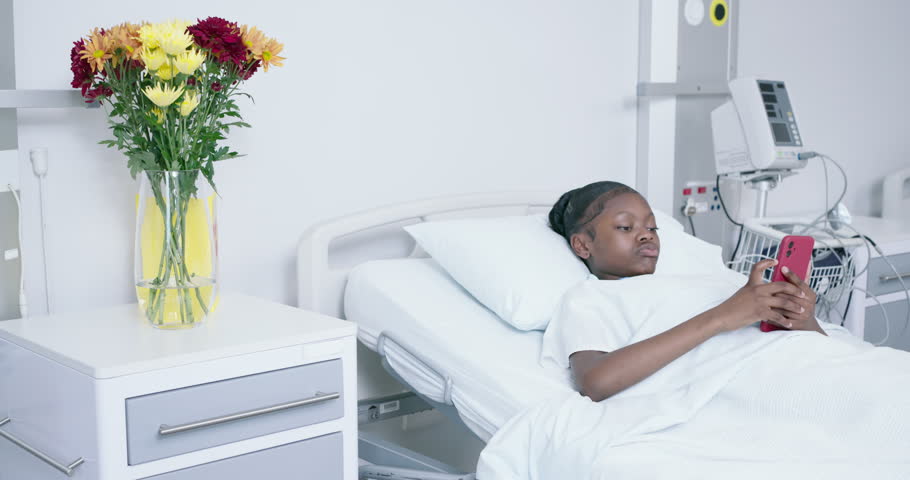 African American teenage patient scrolling red phone in hospital bed lowering phone closing eyes. Clinical, adolescent, device, mobile, bouquet, flowers, monitors