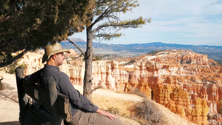 Male tourist wearing cowboy hat sitting on rustic wooden bench under trees, admiring colorful sandstone hoodoos and natural orange rock formations in bryce canyon national park in utah state