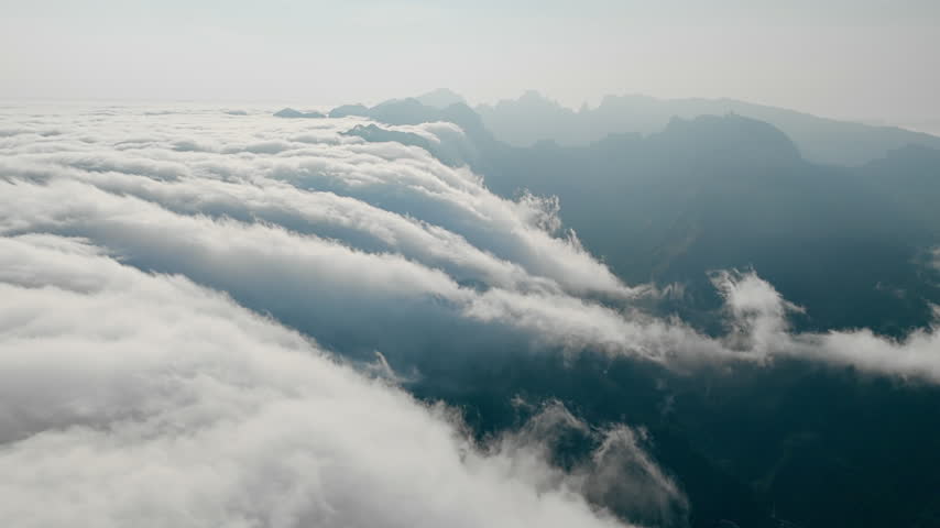 Moving clouds along mountain slopes, aerial view above cloud cover. Rocky peaks rise above cloud layer
