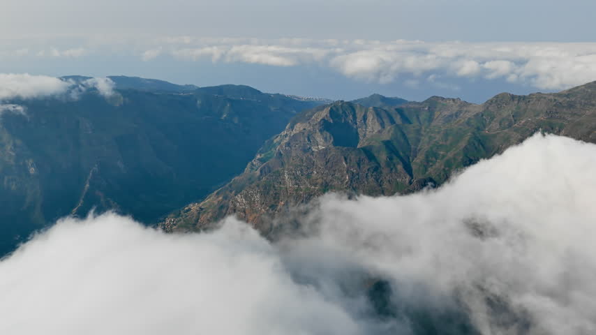 Moving clouds around rugged mountain ridges and steep valleys. Aerial view above low cloud cover.