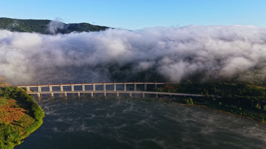 A river flows under a bridge with fog rising from the water. The bridge connects two green areas. Hills are seen in the background against a clear blue sky.
