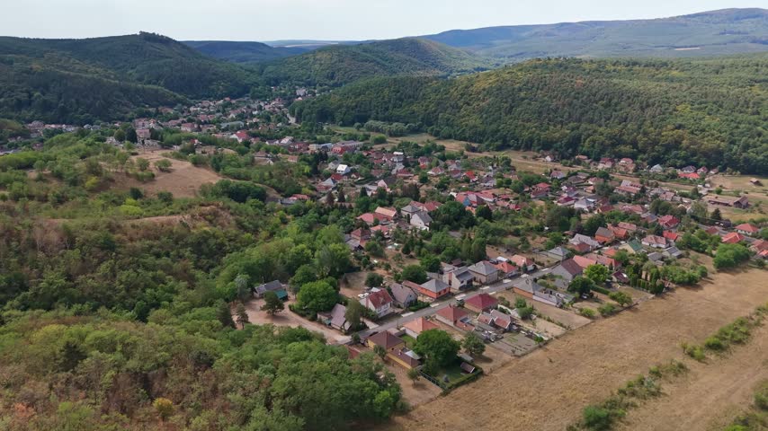 Aerial view of Sirok village in Hungary, showing homes, gardens and cultivated fields in a forested valley surrounded by rolling summer hills.