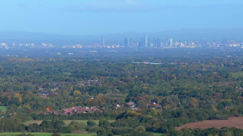 Telephoto view from Alderley Edge across autumn trees and houses of the Cheshire Plain to the Manchester city skyline on the hazy horizon, UK.