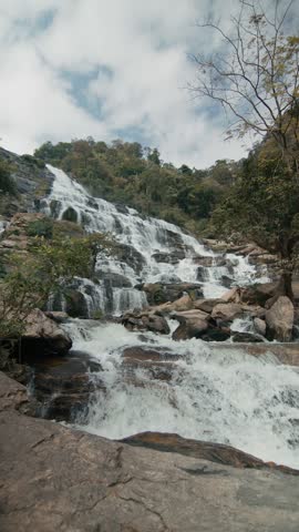 Young woman stands facing cascading mountain waterfall with arms outstretched, slowly lifting hands upward in joyful, freeing gesture, wooded rocky river setting with rushing water, mossy stones, and