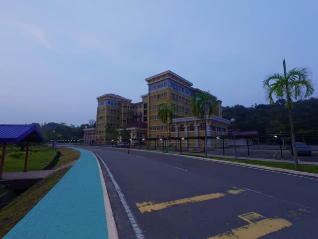 A wide, static establishing shot of a wide campus road with a designated blue bicycle lane curving past modern university buildings under a calm dusk sky.