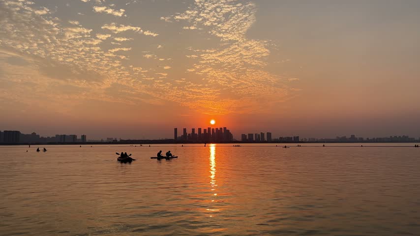 4K static wide shot of a golden sunset over Dushu Lake in Suzhou China. Silhouettes of people on small boats and kayaks traverse shimmering water against a distant city skyline under an orange evening sky.