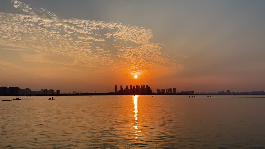 4K static wide shot of a golden sunset over Dushu Lake in Suzhou China. The warm sun sets behind distant urban skyline silhouettes, casting a brilliant orange reflection on rippling water under patterned clouds.