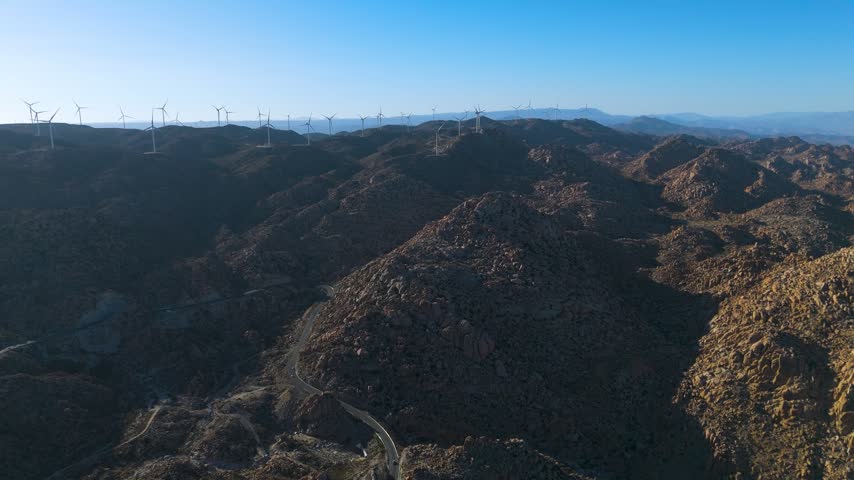 La Rumorosa, Baja California, Mexico, a high aerial view sweeps across rows of granite mountains partly in shadow, their sunlit faces glowing in shades of ochre and rust while the valleys sink into deep shade. A thin road winds through the folds of the terrain in the lower part of the frame, appearing and disappearing between rocky slopes. On the horizon, dozens of white wind turbines line the high plateau of the Sierra de Juarez, with distant blue mountain ranges fading under a clear sky in the late afternoon light, showcasing the vast, remote landscape of northern Baja California.