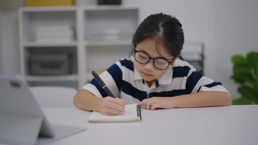 Young girl with glasses writing in notebook focused on homework at a white table with digital device nearby