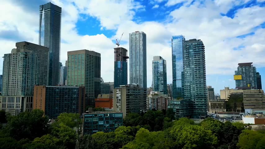 A scenic view of the modern downtown Toronto skyline featuring high-rise residential and commercial skyscrapers, contemporary glass architecture, and lush green trees in the foreground under a beautiful blue sky with white clouds. The urban cityscape showcases Toronto