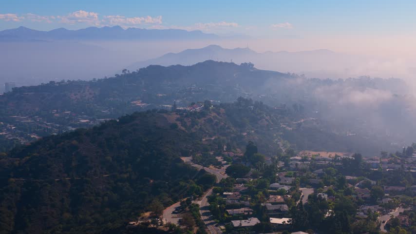 Wooded hills with a winding road and houses below, layered mountain ridges fading into haze and low fog. Radio towers on the hilltop, a dry brown field and clouds in a blue sky. Calm aerial view.