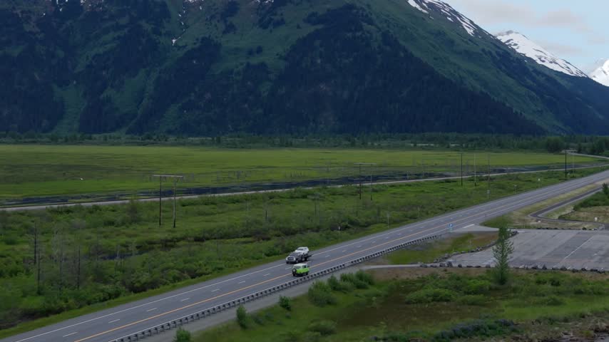 Aerial slow motion descent toward a big parking lot beside the highway with a green van, green wetlands and a mountain slope, Turnagain Arm, Seward Highway, Alaska, USA