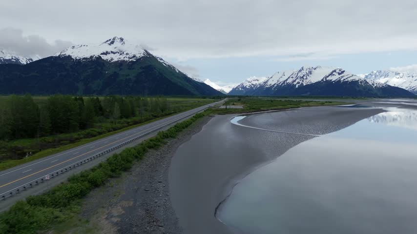 Aerial slow motion forward flight along a highway past tidal flats with sand ripples, a dark car and snow covered mountains, Turnagain Arm, Seward Highway, Alaska, USA