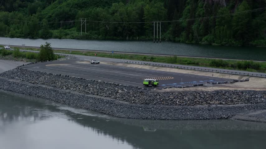 Aerial slow motion forward move over a gravel breakwater toward a roadside parking area with a green van beside calm tidal water, Turnagain Arm, Seward Highway, Alaska, USA