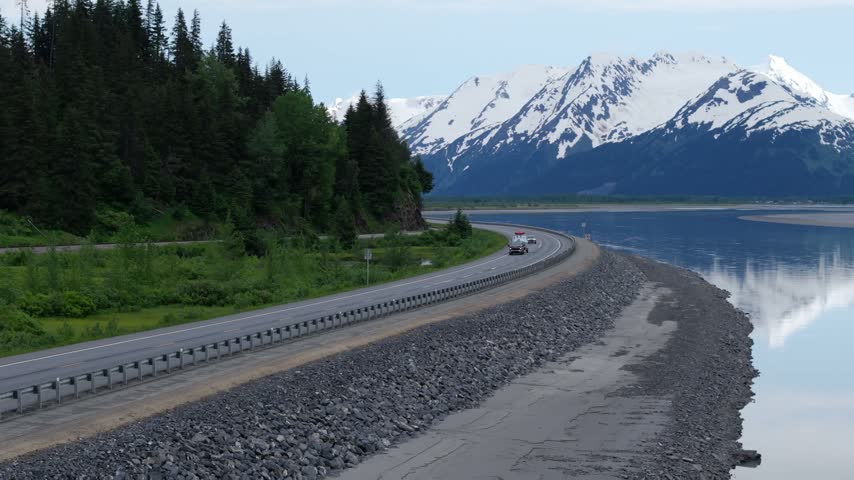 Aerial slow motion forward flight along a curving highway with guardrail, spruce forest, mirror calm water and a snow covered mountain ridge, Turnagain Arm, Seward Highway, Alaska, USA