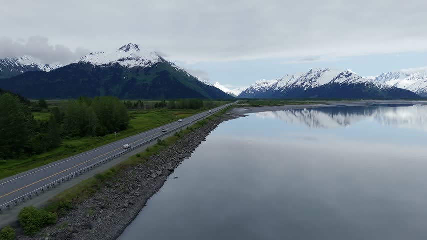 Aerial slow motion forward flight along a highway embankment beside calm water and green lowland with a snow covered peak ahead, Turnagain Arm, Seward Highway, Alaska, USA