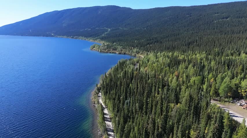 Drone flyover of a vivid blue lake curving around a forested peninsula, with a long mountain ridge and clear sky stretching across the horizon in bright sun