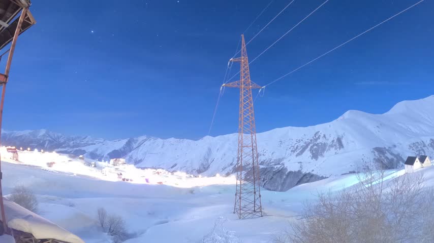 Timelapse view of star trails streaking across a deep blue night sky over illuminated ski resort town lights and snow-covered mountains in Gudauri, Georgia.