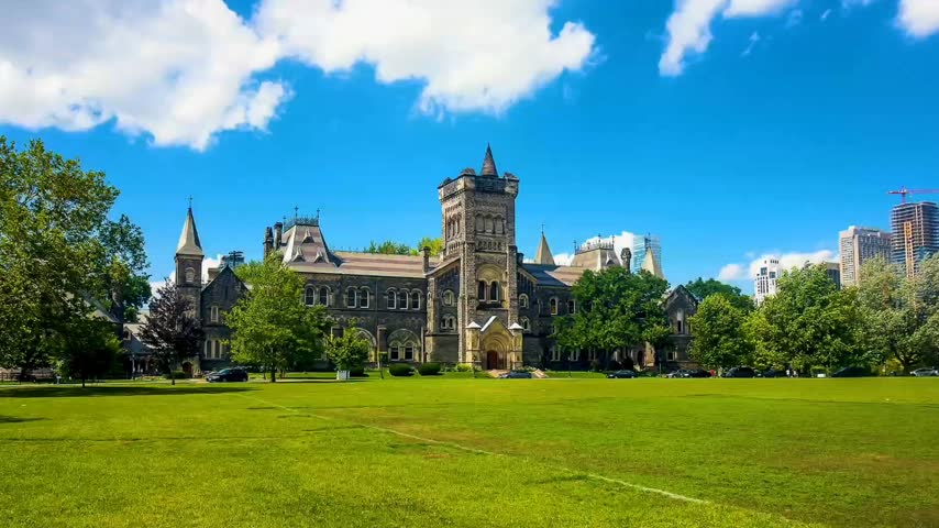 Wide-angle daytime footage of University College at the University of Toronto in Toronto, Ontario, Canada. The historic Gothic Revival building features ornate stone architecture, a prominent central tower, pointed spires, and intricate architectural details. A green lawn, mature trees, parked cars, and a bright blue sky create a picturesque university campus setting. Ideal for educational content, Canadian travel documentaries, university and academic themes, historic architecture, tourism promotions, and establishing shots of Toronto landmarks.