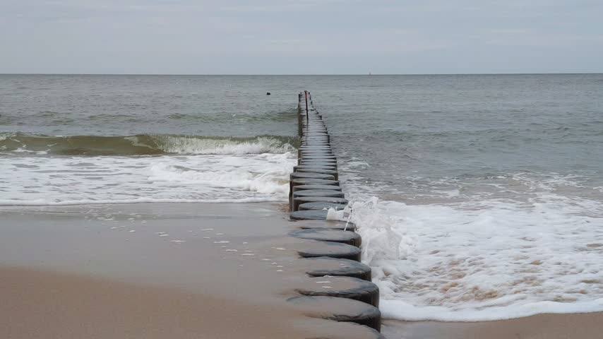 Close-up of stormy foamy coastal sea waves breaking on old wooden breakwater and wet sand on empty sandy beach seashore under cloudy sky. No people serene seaside landscape. Natural sound of surf.