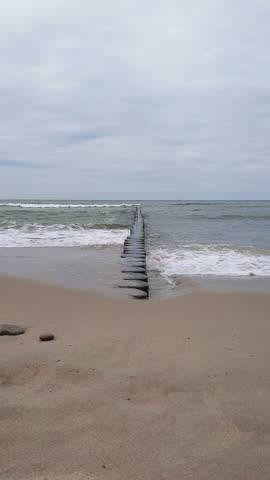 Vertical shot of stormy foamy coastal sea waves breaking on old wooden groyne and wet sand on empty sandy beach seashore under overcast sky. No people serene seaside landscape. Natural sound of surf.