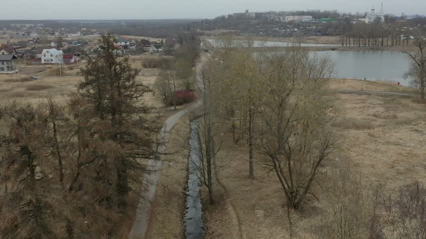 Rural spring landscape featuring tall trees, a narrow river, a lake, and distant houses under an overcast sky.