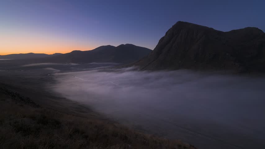 Low fog drifts beneath dark mountain slopes in Glen Coe, Scotland, as a warm dawn glow appears along the horizon.