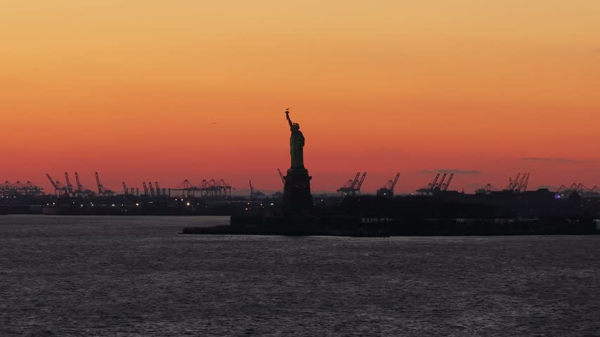 Aerial telephoto view of the Statue of Liberty silhouette against an orange sunset sky with port cranes behind, New York Harbor, New York City, USA
