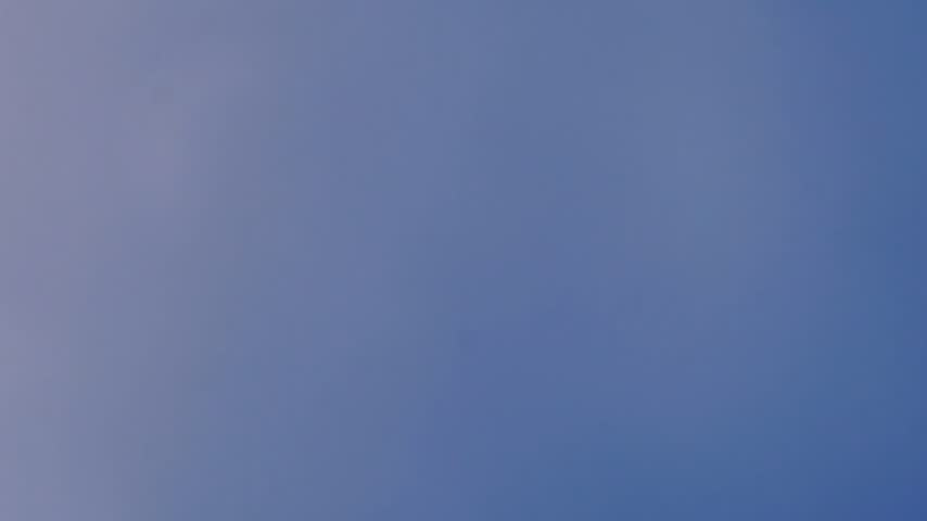 A stunning close-up view of a half moon moving across a bright blue daytime sky, accompanied by soft, passing clouds.