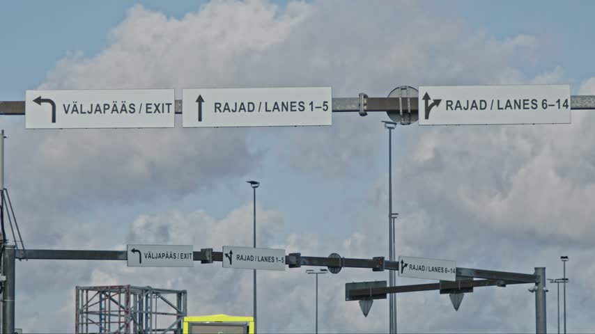 Directional road signs showing exit and numbered vehicle lanes at Tallinn passenger ferry terminal in Estonia. Static 4K footage of port infrastructure with clouds moving across the blue summer sky.