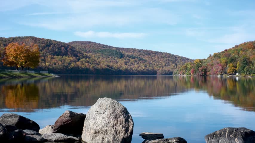 Fall foliage reflects on the calm water of a scenic New England lake with foreground rocks.