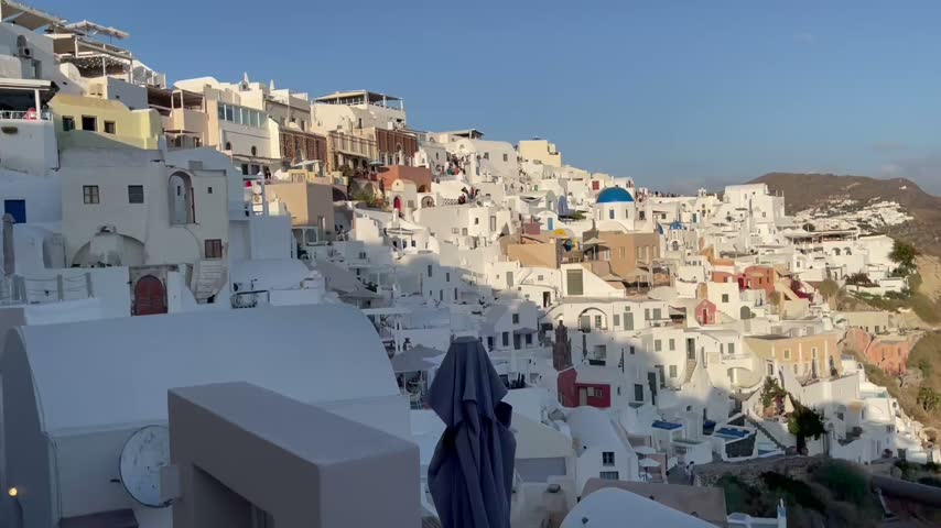 Whitewashed buildings of Oia cling to the volcanic caldera of Santorini, Greece. Scenic Mediterranean island footage featuring cliffside architecture and coastal views.