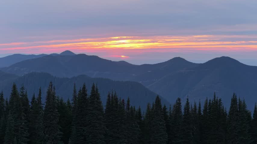 Mountain range sunset illuminates evergreen treetops, layered ridges, and wispy clouds as dusk settles over Chilliwack, British Columbia, Canada.