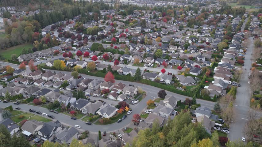 Aerial drone glides over a suburban neighborhood in Chilliwack, British Columbia, revealing homes, streets, colorful fall trees, and forested hills under a hazy sky.