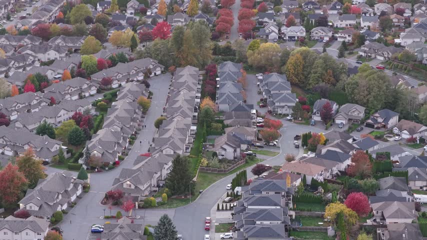 Aerial view slowly pans across a Chilliwack suburban neighborhood in British Columbia, showing homes, curving streets, and colorful autumn trees in soft daylight.