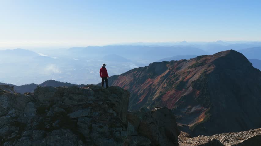 Hiker stands on a rocky mountain peak, views the alpine landscape, then removes a backpack in an aerial view above Chilliwack, British Columbia.