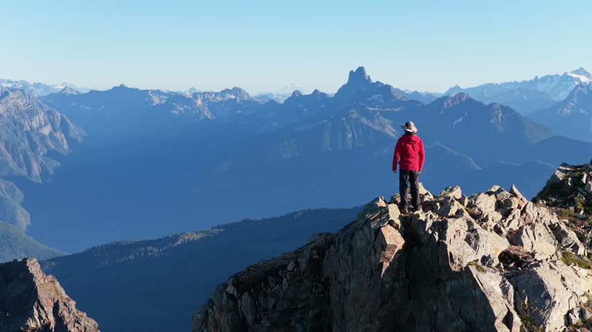 Hiker stands on a rocky mountain summit and admires panoramic mountain views as an aerial drone zooms out across Chilliwack, British Columbia, Canada.