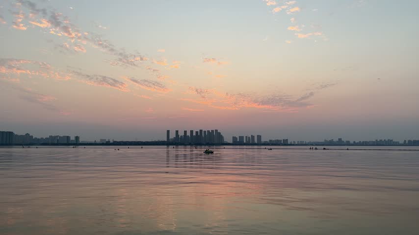 4K static wide shot of Dushu Lake in Suzhou during twilight. A bird flies across the frame above calm water reflecting the soft pink sunset sky and distant city skyline.