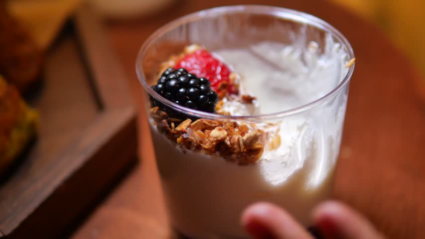 Close up shot of hand using spoon to scoop delicious yogurt parfait with blackberries strawberries and crunchy granola from small plastic cup on wooden table.