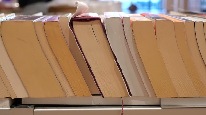 Rows of various paperback books lined up on wooden shelf inside library for research and education. Stack of books organized for readers and students in quiet indoor atmosphere.