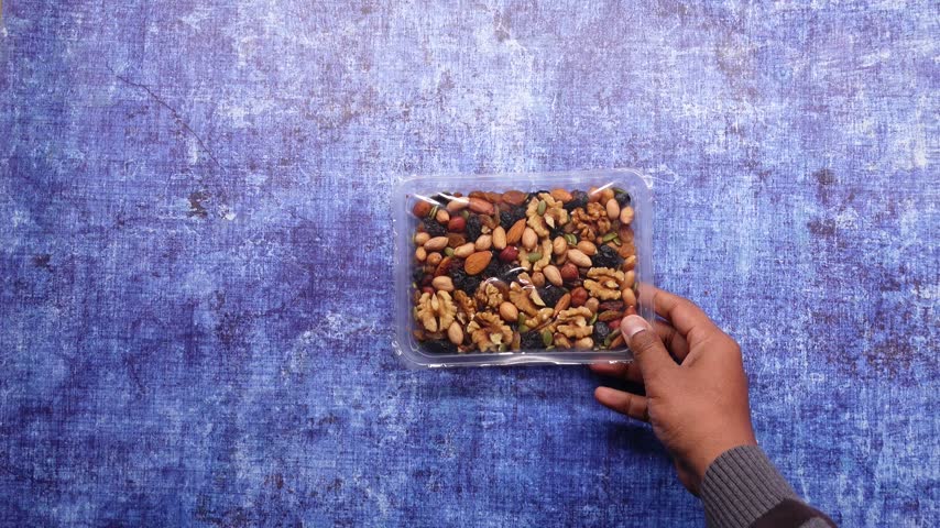 Person holding plastic container filled with assortment of mixed nuts and dried fruits including almonds walnuts cashews and raisins on textured blue background close up shot.