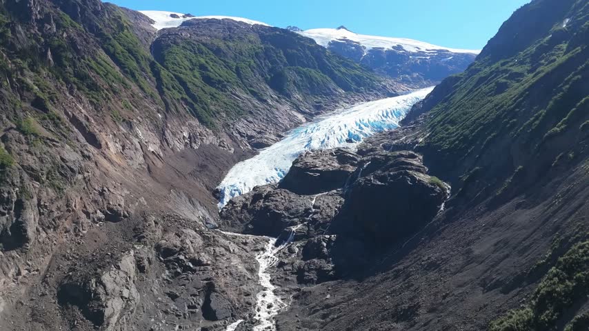Drone flyover of a glacier tongue descending between steep rocky slopes with a meltwater stream and snow-capped peaks under a blue sky near Stewart, British Columbia, Canada