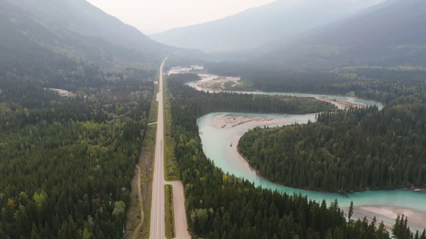 Drone shot of a highway running alongside a winding turquoise river and sandy gravel bars in a forested mountain valley near Jasper, Alberta, Canada