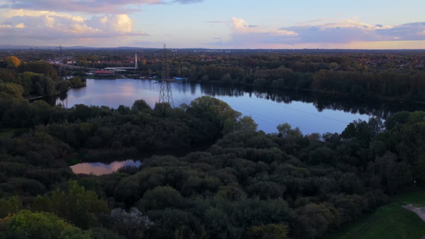 Drone view of Sale Water Park lake at dusk with an electricity pylon, a footbridge over the M60 and woodland at Stretford Ees under a pastel sky.