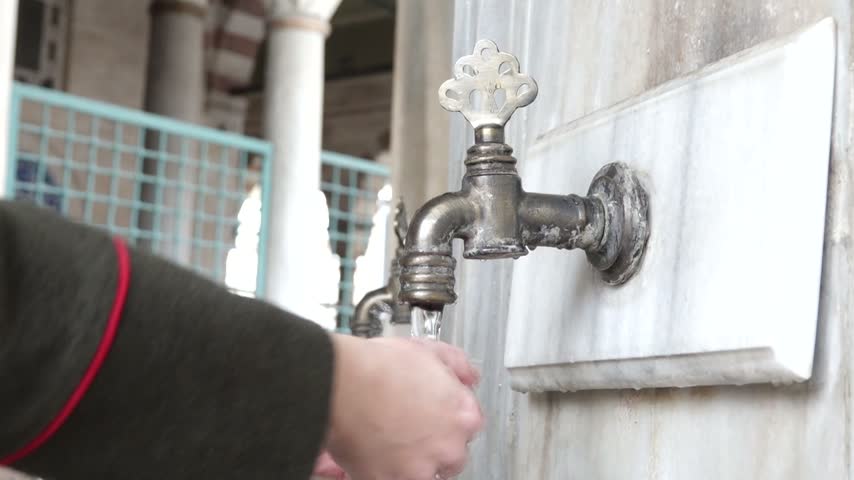 Close-up of a woman washing her hands under a traditional Ottoman fountain tap in the courtyard of a historic mosque in Istanbul, Turkey.