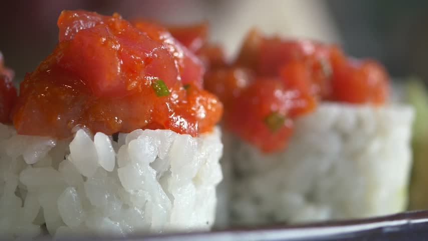 Close up shot of fresh spicy tuna sushi rolls with raw fish topping served on dark plate. Culinary footage showcasing authentic Japanese dish with white rice and red tuna topping.